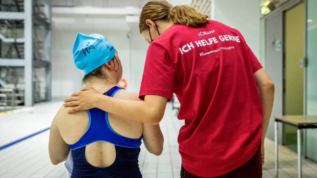 Ein Person im rotem T-Shirt mit dem Aufdruck "Ich helfe gern" beugt sich über eine junge Frau im blauen Badeanzug und einer Badekappe.