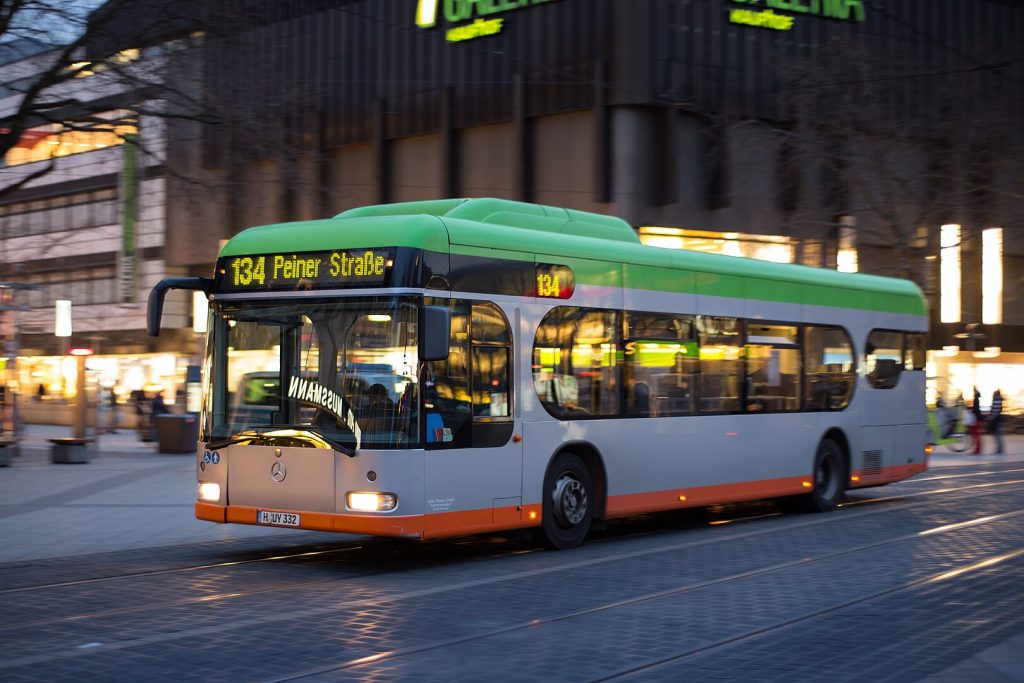 Bus von UESTRA der auf einer Straße fährt. Der Bus ist grün, grau und orange und trägt die Nummer 134 zur Peiner Straße. Der Bus fährt in der Dämmerung und seine Lichter sind an.