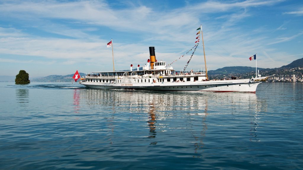 Weißes Schiff fährt auf Wasser mit Sonnenschein. Vorne trägt das Schiff eine französische Flagge, hinten eine schweizerische Flagge.