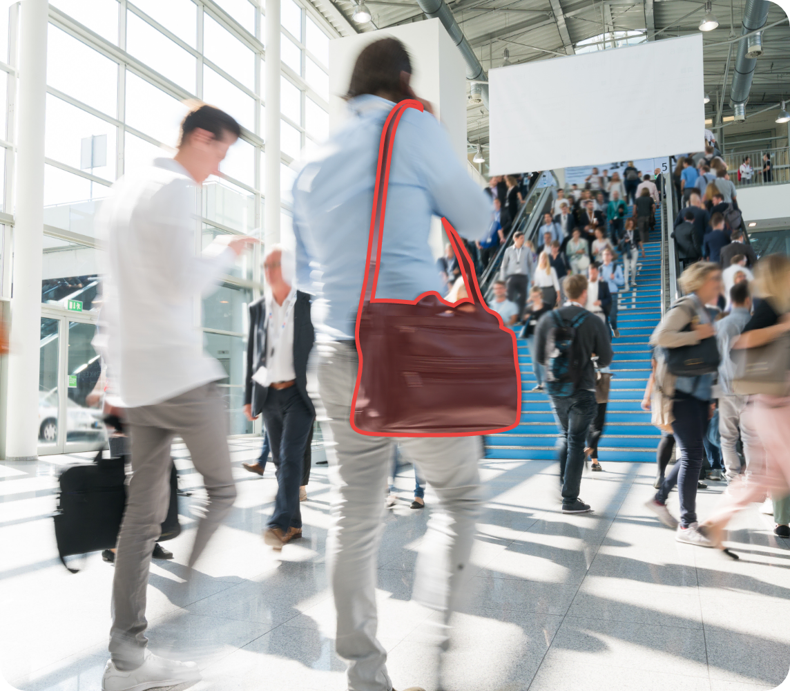 Messehalle in der Menschen gehen in Business-Kleidung. Zwei Männer von hinten stehen im Fokus, bei einem ist die Tasche rot hervorgehoben.